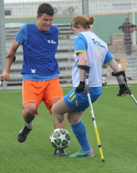 A woman on crutches and a coach play soccer together on a turf field