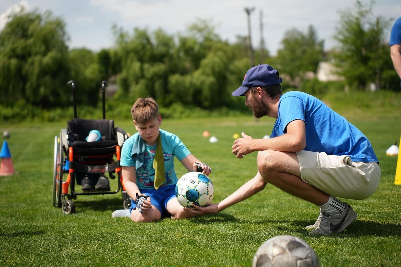 A coach kneels to pass a ball to a boy in a wheelchair on the pitch